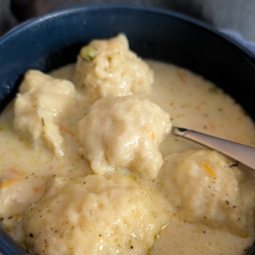 Creamy chicken and dumplings in a bowl with soft doughy dumplings, vegetables, and a spoon in a thick, comforting broth.
