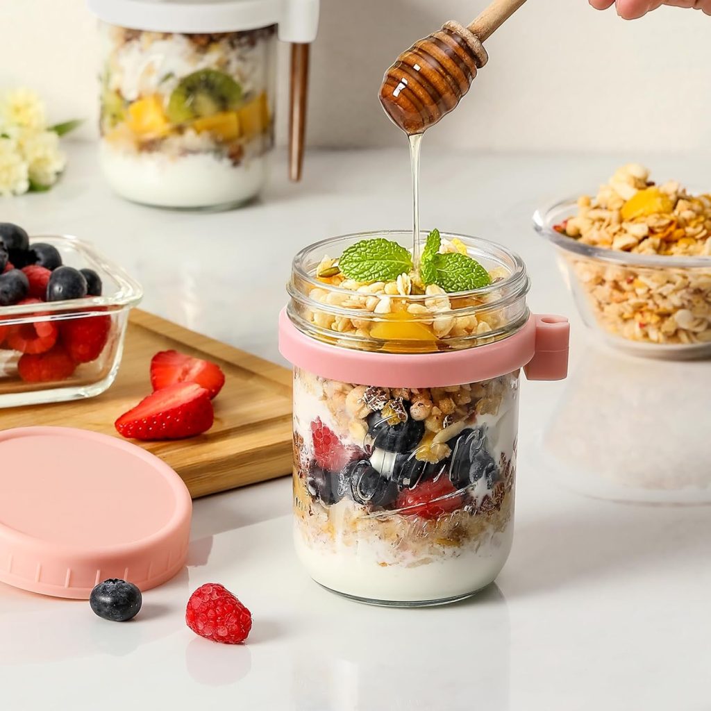 Jar layered with yogurt, granola, and fresh berries while honey is drizzled on top, surrounded by bowls of fruit and granola on a white surface.
