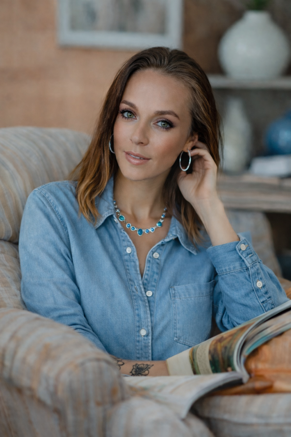 Person sitting in a striped armchair wearing a light denim shirt, hoop earrings, and a blue gemstone necklace while holding an open magazine in a cozy indoor space.