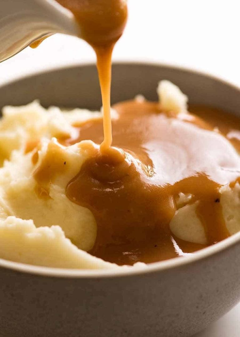 Close‑up of creamy mashed potatoes being topped with warm brown gravy poured from a white container.