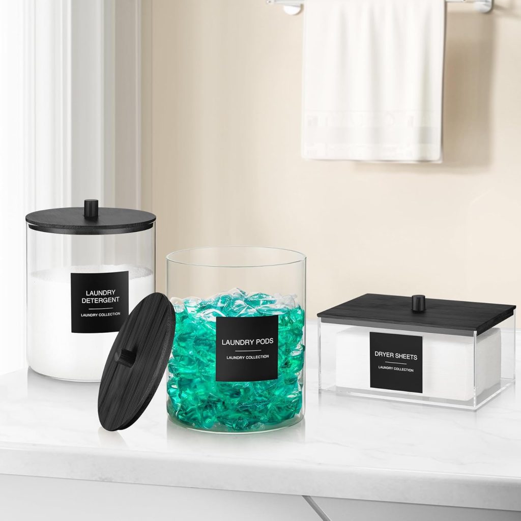 Organized laundry counter with labeled clear containers holding detergent powder, laundry pods, and dryer sheets on a white surface.