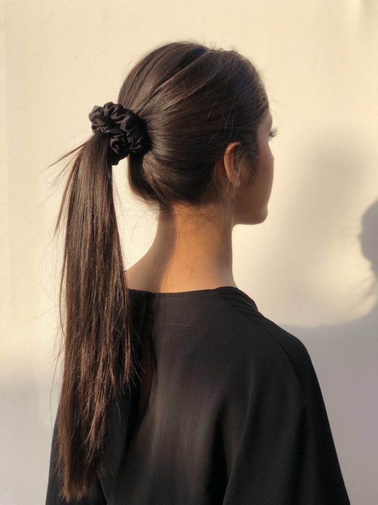 Back view of long dark hair tied in a ponytail with a black scrunchie, worn with a black top against a light background in natural sunlight.