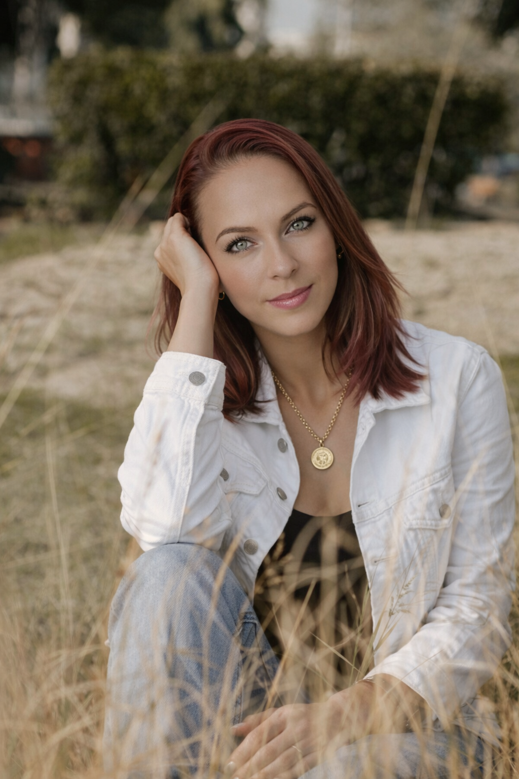 Person wearing a white denim jacket, black top, blue jeans, and a gold pendant necklace sitting outdoors on grass with greenery in the background.