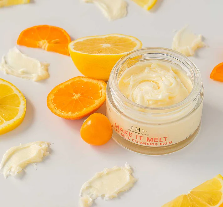 A jar of Make It Melt Silky Milk Cleansing Balm surrounded by sliced citrus fruits and small dollops of balm on a white surface.