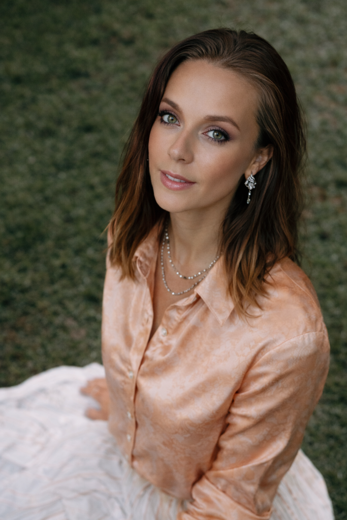 Person sitting outdoors on grass wearing a peach satin blouse, white skirt, pearl necklaces, and earrings, looking upward in soft natural light.