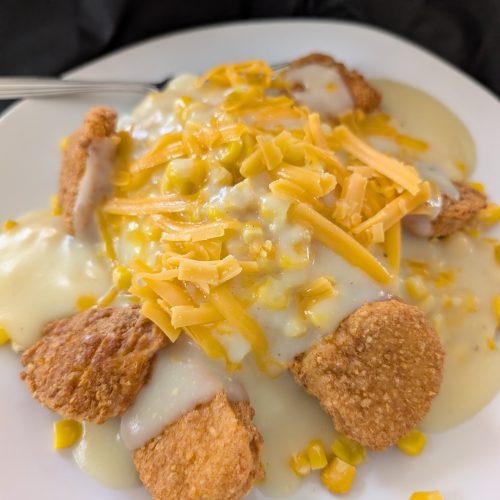 Plate of crispy chicken nuggets topped with creamy white gravy, sweet corn, and melted cheddar cheese, served as a cozy comfort‑food bowl.