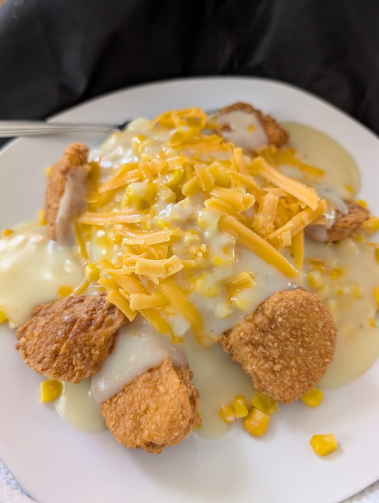 Plate of crispy chicken nuggets topped with creamy white gravy, sweet corn, and melted cheddar cheese, served as a cozy comfort‑food bowl.