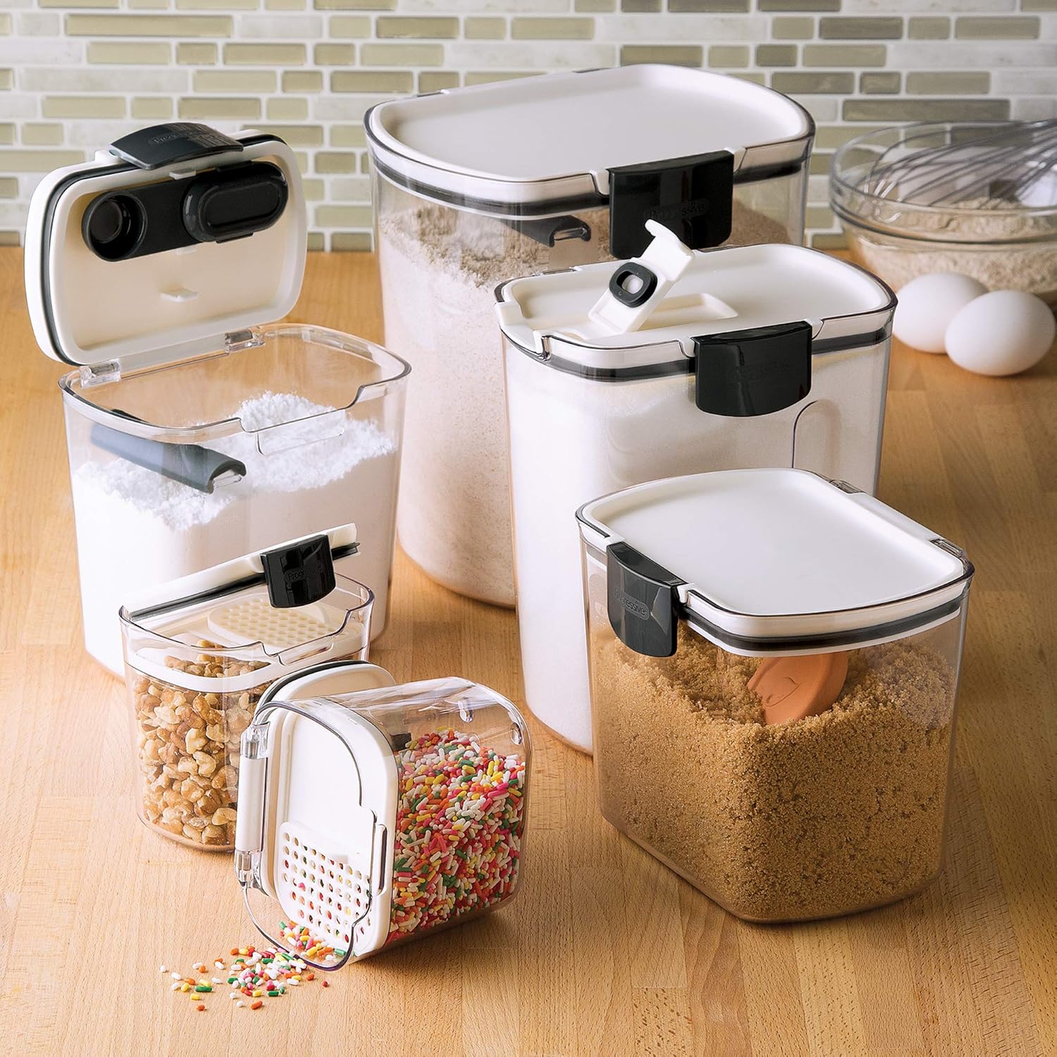 Clear food storage containers with white lids holding powdered sugar, flour, brown sugar, nuts, and sprinkles arranged on a wooden kitchen countertop.