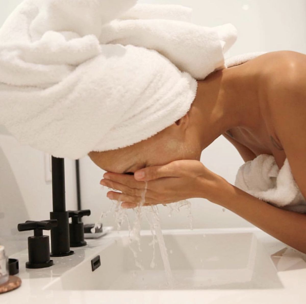 Person washing their face at a bathroom sink with natural light, wearing a towel around their head and body, water splashing from their hands.