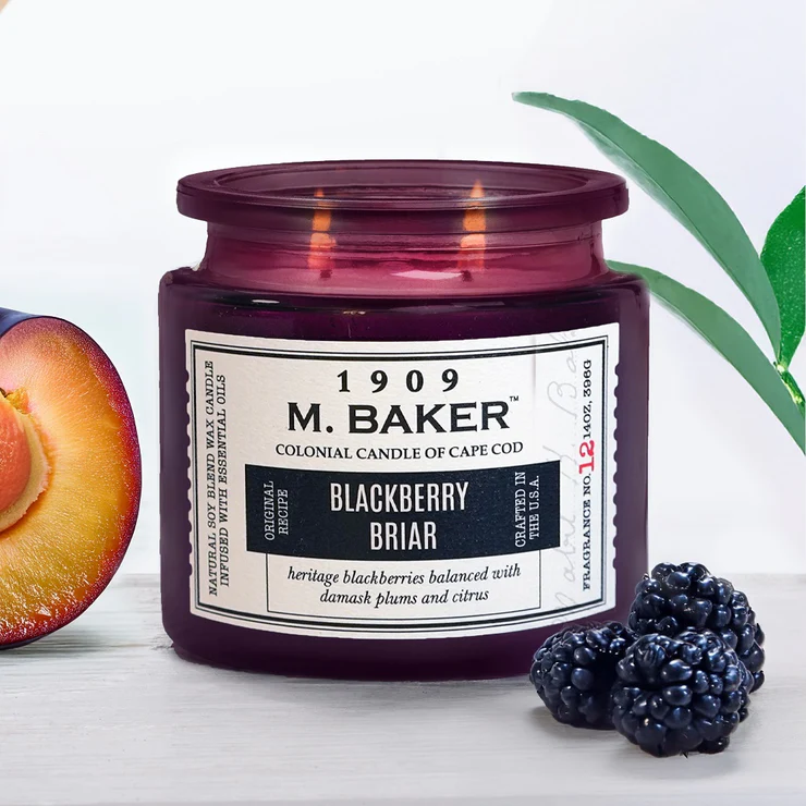 A lit purple glass jar candle labeled “1909 M. Baker Colonial Candle – Blackberry Briar” sits beside fresh blackberries, a halved plum, and a green leaf. The warm glow highlights the candle’s rich berry, plum, and citrus scent profile in a moody, artisanal setting.
