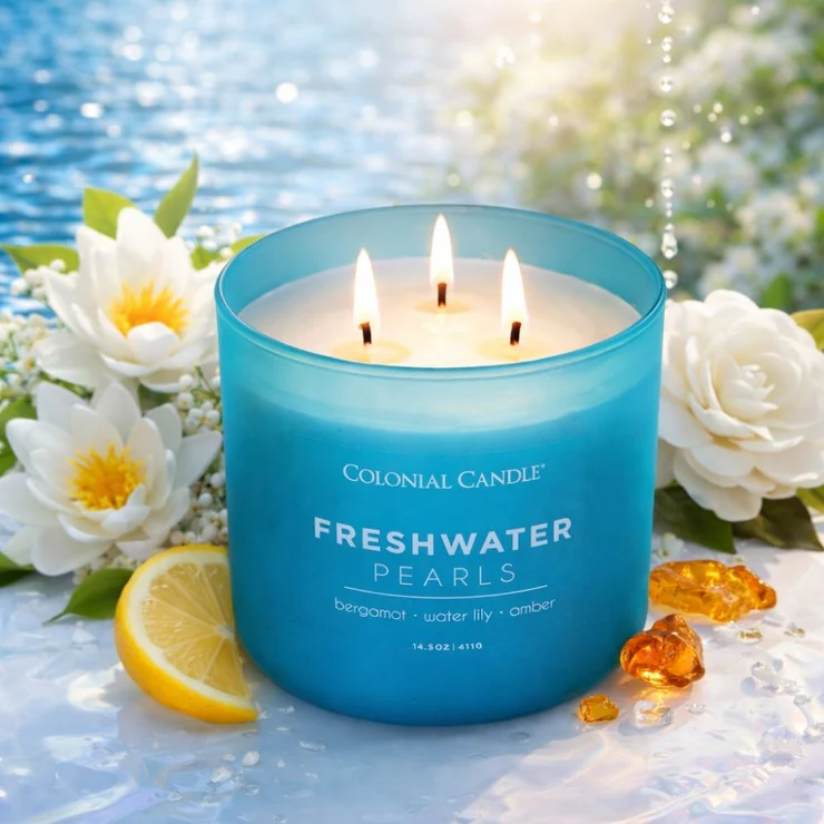 A lit three‑wick blue candle labeled “Colonial Candle Freshwater Pearls” sits surrounded by white water lilies, a lemon slice, amber resin pieces, and small white blossoms. Soft sunlight reflects off a calm water backdrop, creating a fresh, serene, spa‑like atmosphere that highlights the candle’s bergamot, water lily, and amber scent profile.