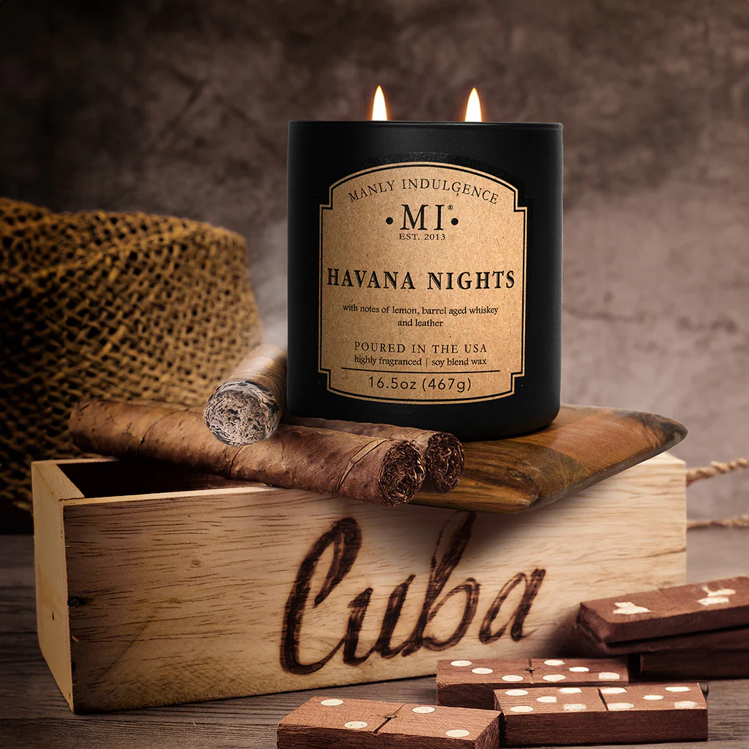 A lit black “Havana Nights” candle sits on a wooden surface surrounded by cigars, a wooden Cuba box, and scattered wooden domino pieces. The warm glow and rustic textures create a rich, vintage, masculine atmosphere that highlights the candle’s whiskey, leather, and citrus notes.