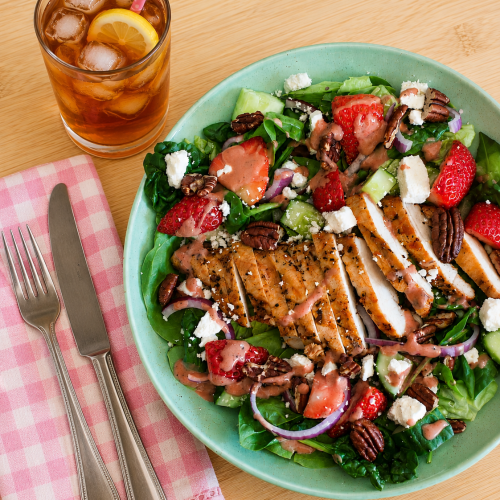 Colorful spinach salad with grilled chicken, strawberries, cucumbers, red onions, pecans, feta, and pink dressing served with iced tea on a wooden table.