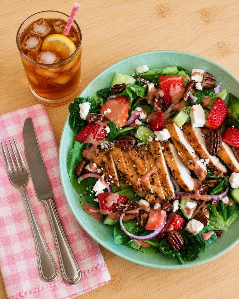 Colorful spinach salad with grilled chicken, strawberries, cucumbers, red onions, pecans, feta, and pink dressing served with iced tea on a wooden table.