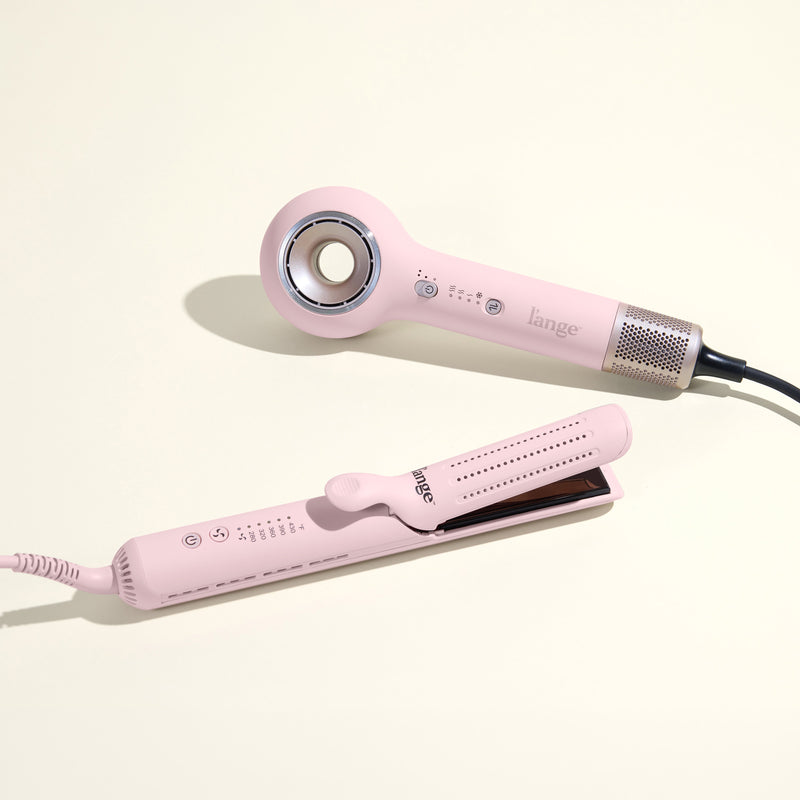Two pink L’ange hair styling tools—a modern hollow‑center hair dryer and a sleek flat iron—placed on a cream surface with visible control buttons and power cords.