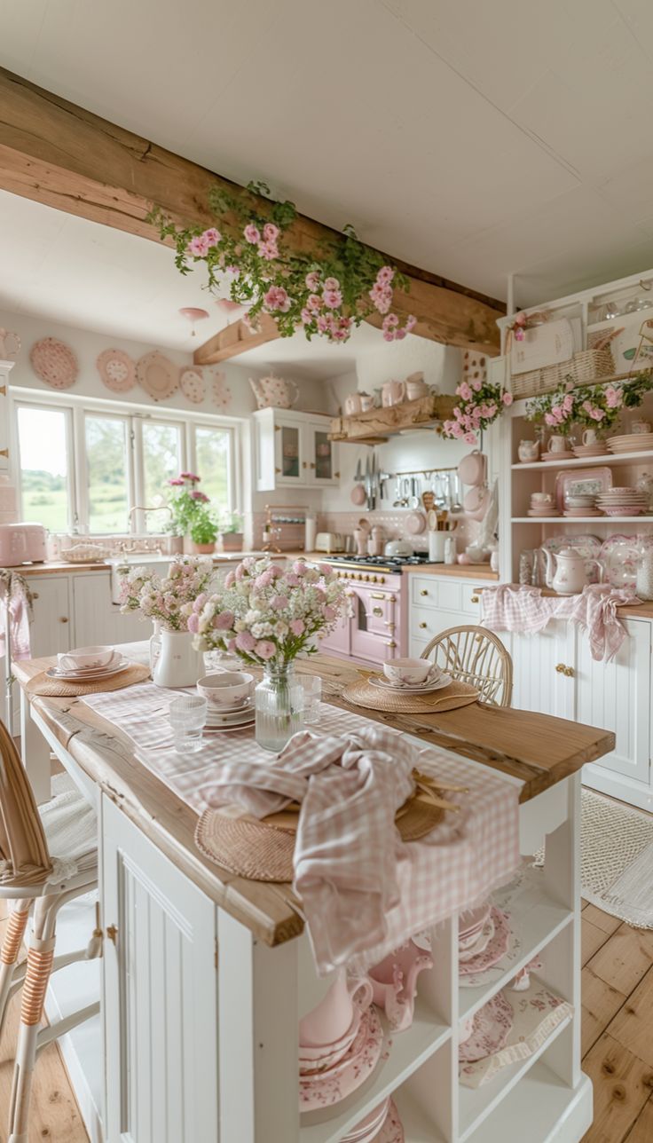 A soft pink vintage-style kitchen with white cabinetry, wooden countertops, floral garlands on ceiling beams, a pink stove with brass accents, and a dining table set with pink checkered linens, woven placemats, glassware, and pink rose centerpieces.