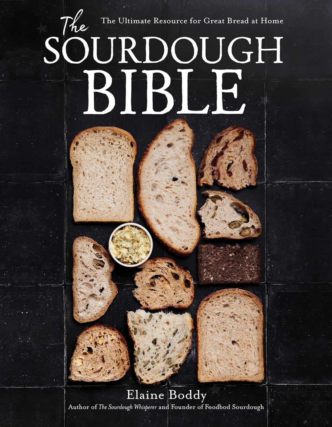 The Sourdough Bible cookbook displayed on a white wooden stand, featuring a rustic loaf of sourdough bread on the cover with baking ingredients in the background.